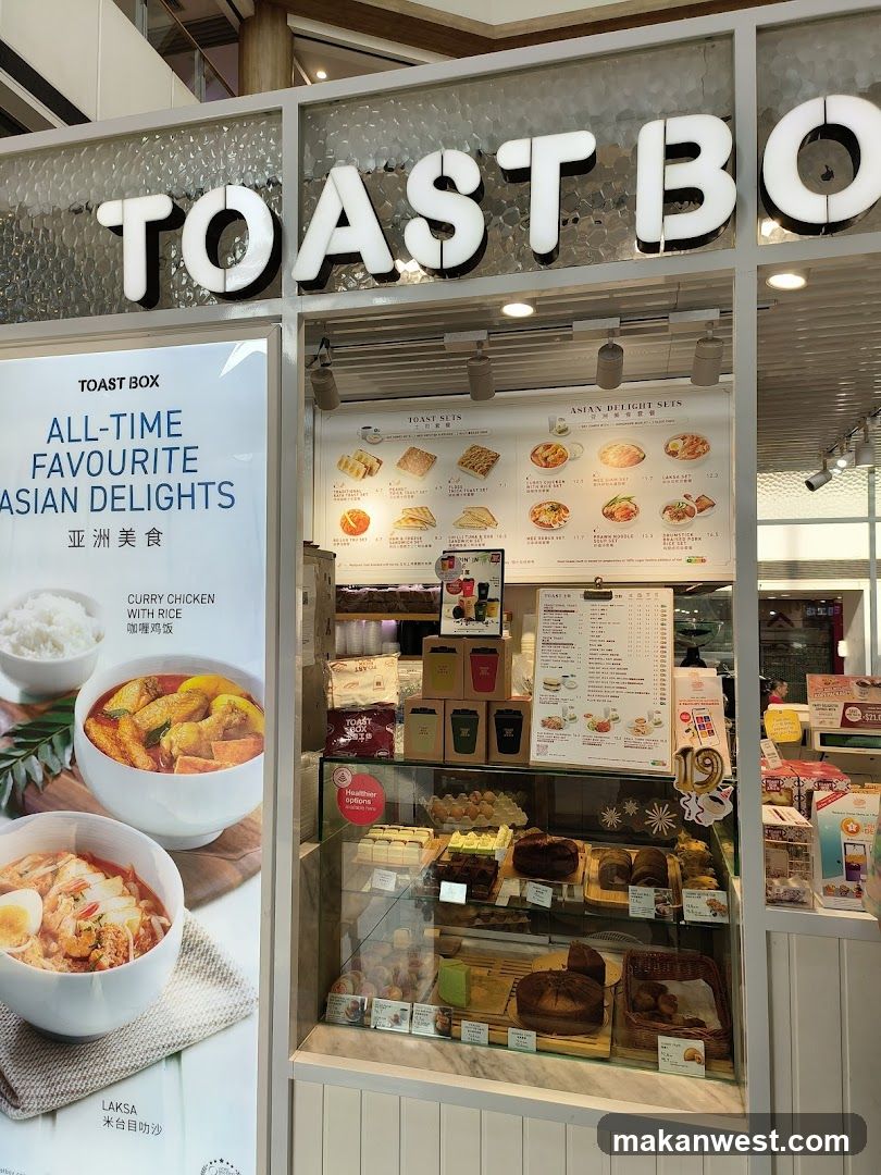 A brightly lit shop front with large illuminated signage reading 'TOAST BO'. A glass display case showcases various baked goods and pastries, including breads and cakes. To the left, a poster advertises 'ALL-TIME FAVOURITE ASIAN DELIGHTS' featuring images of curry chicken with rice and laksa. Inside, menu boards list Asian delights such as toast meals and rice dishes.
