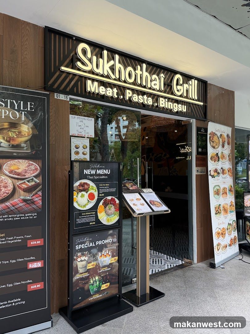 Photo of the storefront facade showing illuminated signage reading 'Sukhothai Grill' with subtitle 'Meat . Pasta . Bingsu'. A glass entrance is visible, flanked by promotional posters and menu boards displaying food items like Thai specialties and special promos.