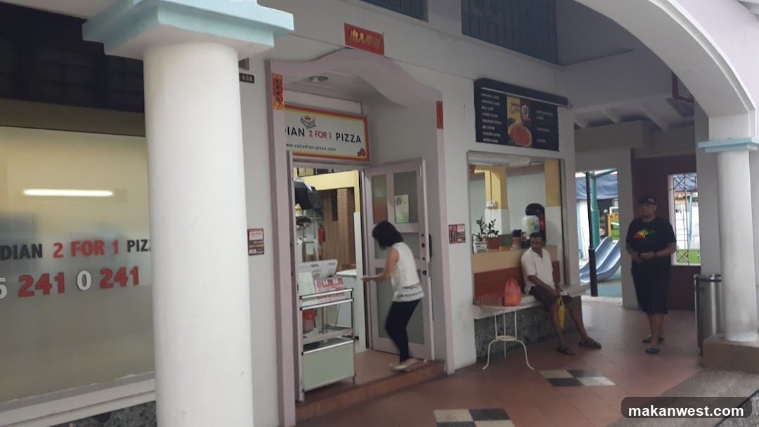 A storefront with a white facade and large columns. A woman is entering the shop through glass doors. To the right, two men sit on a bench near a menu board displaying pizza options. The building features signage reading 'INDIAN 2 FOR 1 PIZZA' above the entrance and on the window.