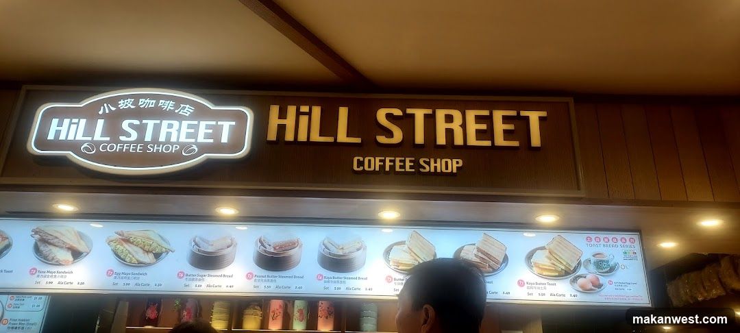 A brightly lit storefront sign displaying 'HILL STREET COFFEE SHOP' in English and Chinese characters, mounted above a food display counter with illuminated menu boards showing various sandwich items.