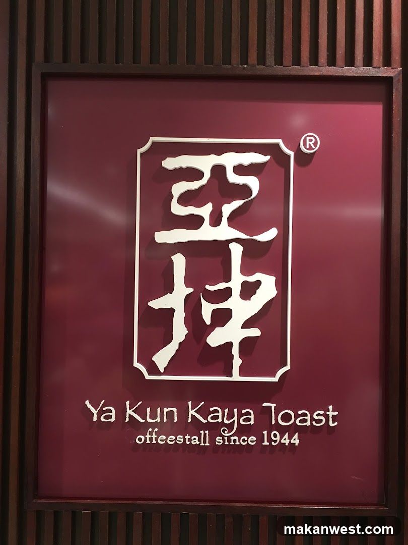 A close-up photo of a shop sign featuring the Chinese characters '亞坤' (Ya Kun) in white on a maroon background, with English text below reading 'Ya Kun Kaya Toast' and 'offeestall since 1944'. The sign is mounted on a wooden slatted wall.