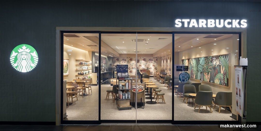 A modern Starbucks storefront with large glass windows and doors, illuminated signage above the entrance, and a glowing green siren logo on the left wall. Inside, wooden tables and chairs are arranged in an open layout, with decorative wall art featuring tropical foliage patterns. A promotional stand is visible near the entrance.
