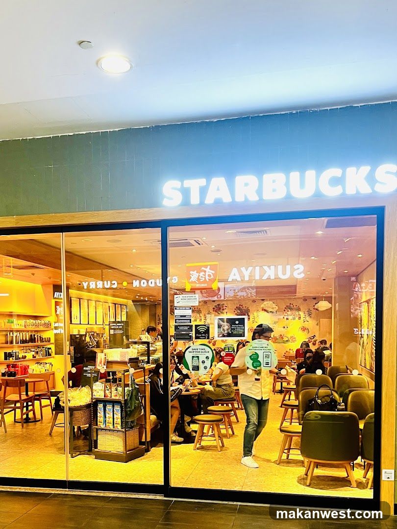 The image shows the exterior of a Starbucks store with its illuminated sign clearly visible above the entrance. The storefront features large glass windows and doors, allowing a view into the interior where customers are seated at tables. Inside, there is a warm lighting ambiance with wooden furniture and various menu boards and promotional materials displayed on the walls.