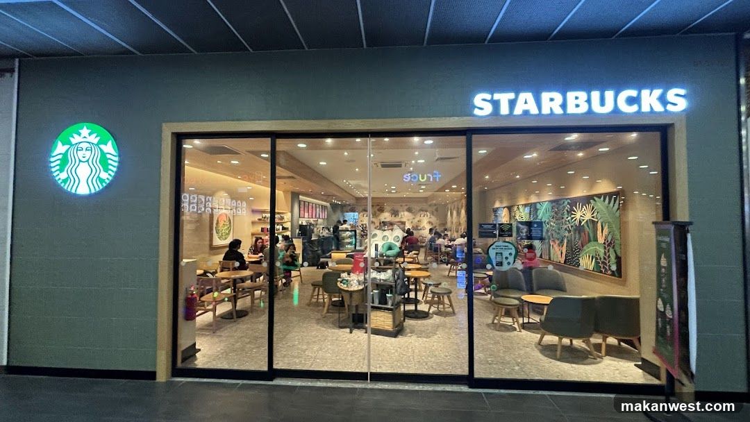 A Starbucks storefront with illuminated signage and logo on the exterior wall. Large glass windows reveal an interior seating area with customers dining, tables, chairs, and decorative wall art featuring tropical designs.