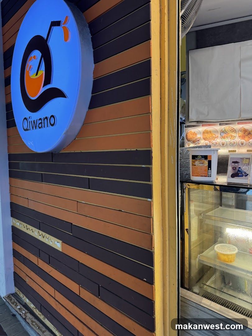 A shop front with a blue circular sign displaying the logo 'Qiwano' featuring an orange and black design, mounted on a wall with horizontal brown and dark wood slats. To the right, there is a glass display case containing food items and a menu board showing various dishes with prices.
