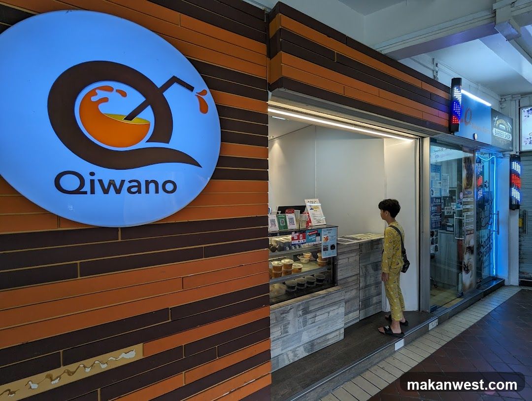 A shop front with a large circular sign displaying the logo and name 'Qiwano' on an orange-brown brick wall. The entrance is open, revealing a display counter inside where a person in yellow clothing stands near the glass door.