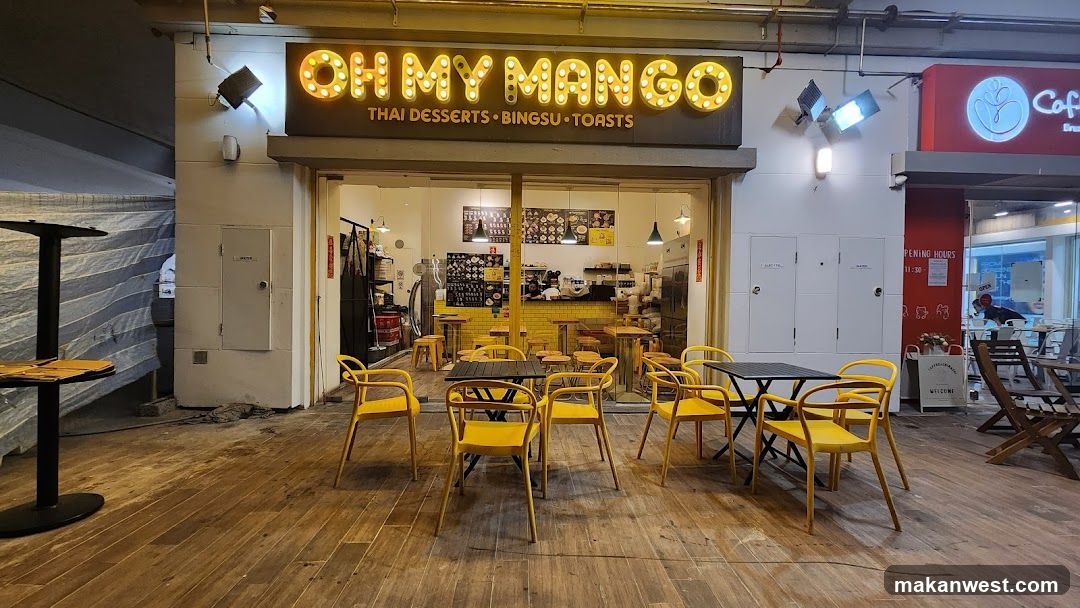 The image shows the storefront of 'OH MY MANGO', a Thai dessert shop with illuminated signage reading 'THAI DESSERTS • BINGSU • TOASTS'. The entrance is open, revealing an interior with yellow walls and tables. Outside seating includes several yellow chairs around black tables on a wooden floor. To the right, another shop sign partially visible reads 'Café'. No menu boards or price lists are clearly visible in the exterior view.