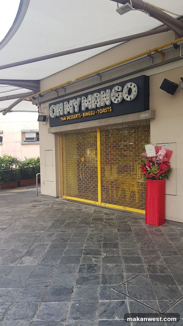 The image shows the exterior of a shop named 'OH MY MANGO' with signage indicating it serves Thai desserts, bingsu, and toasts. The storefront is closed with yellow metal shutters down. A red planter with flowers sits beside the entrance.