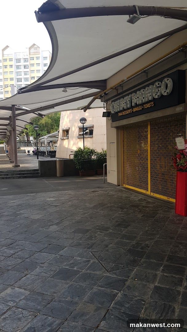 A shop front with a sign reading 'SHINY MANGO' and 'PAW DESSERTS, SINGSU TOASTS'. The storefront has yellow shutters that are closed. There is a red flower pot on the right side of the entrance. A large white awning extends over the sidewalk area.