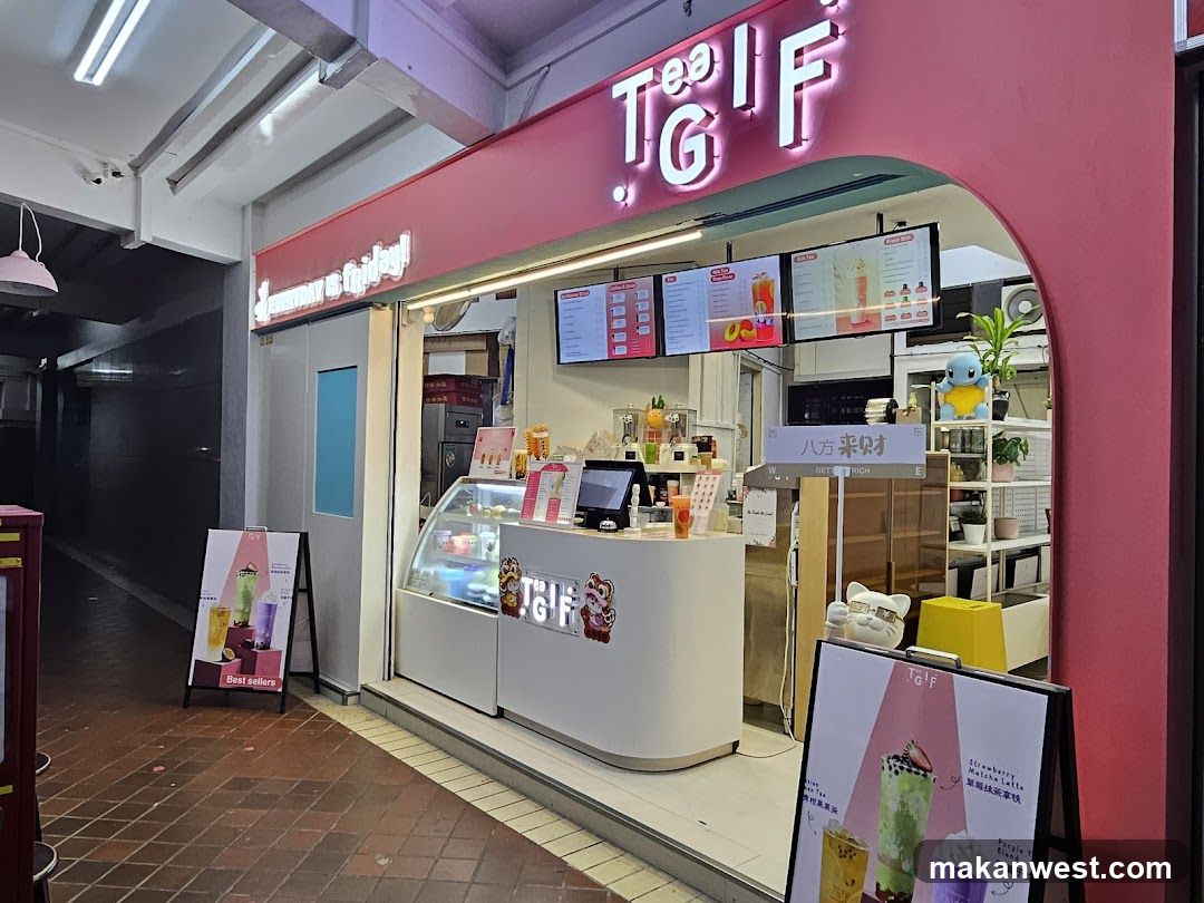 The image shows the storefront of a shop named TGI F, with illuminated signage on the building facade. The entrance features a curved glass display case and menu boards inside. A signboard outside advertises strawberry milk tea.