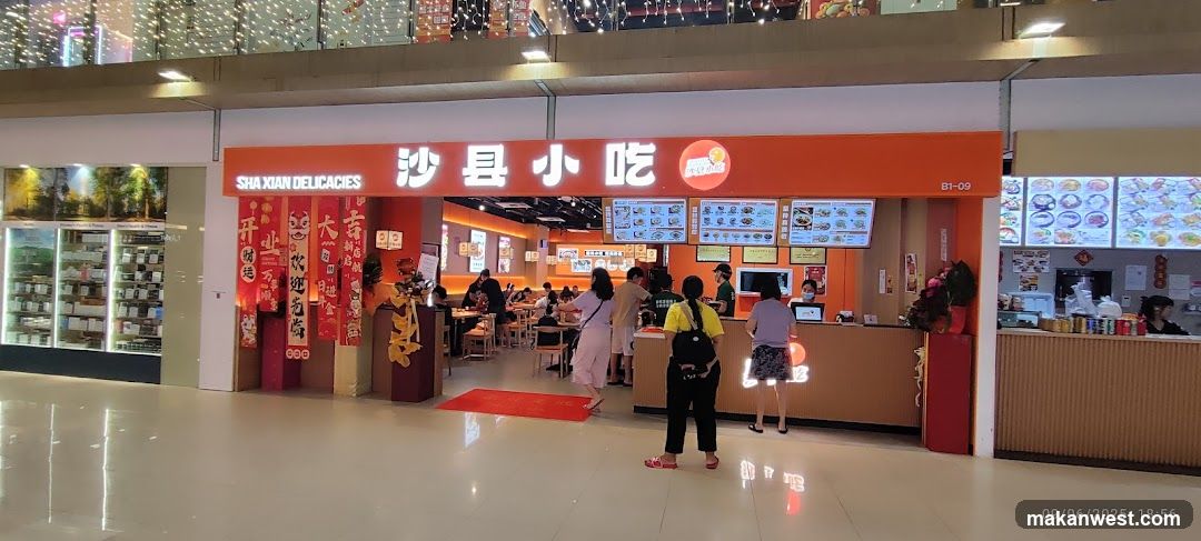 A brightly lit storefront with an orange facade displaying 'SHA XIAN DELICACIES' and Chinese characters '沙县小吃'. The entrance features red banners with festive decorations, including a golden dragon figure. Inside, customers are seated at tables, while others queue near the counter. Menu boards are visible above the service area.