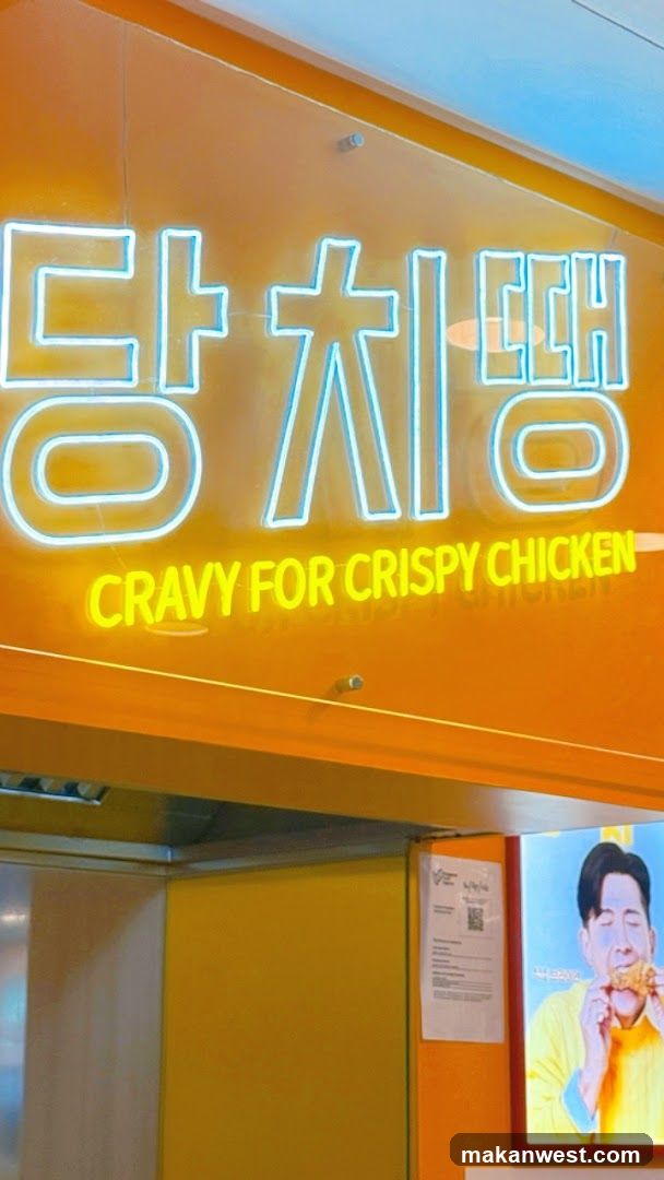 A brightly lit storefront featuring a large orange sign with neon-style Korean characters and the English phrase 'CRAVY FOR CRISPY CHICKEN' in yellow below. Below the main sign, part of an advertisement or poster is visible showing a person eating fried chicken.