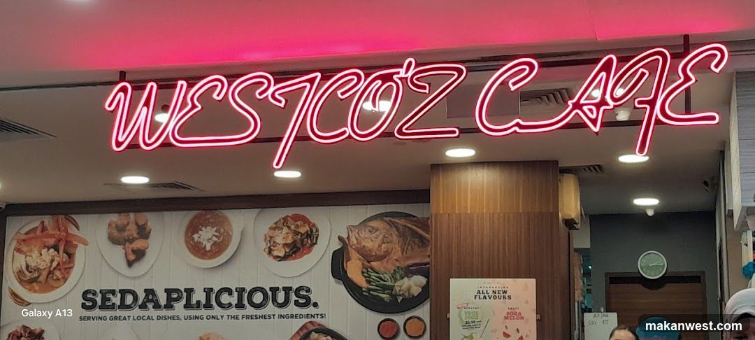 A photo of the physical storefront facade featuring a neon sign reading 'WESTCOZ CAFE' in red cursive letters above the entrance. Below, there is a large wall display showcasing various food items with the slogan 'SEDAPLICIOUS.' and a smaller poster advertising new flavors.