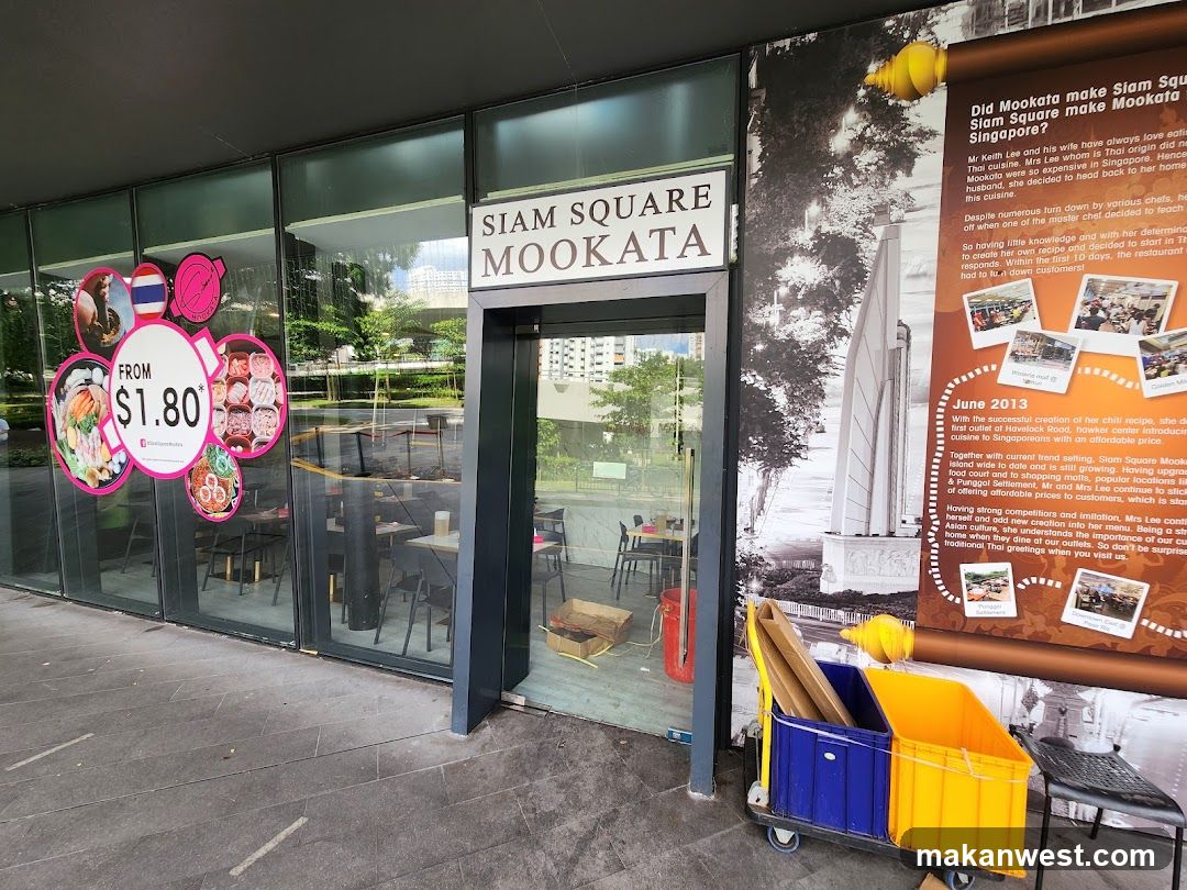 Exterior storefront of Siam Square Mookata, featuring a glass entrance with signage above the door. Large window displays colorful food-themed graphics and promotional pricing (from $1.80). A large mural on the wall to the right depicts historical imagery and text about Mookata culture in Singapore. Yellow and blue storage bins are visible near the entrance.