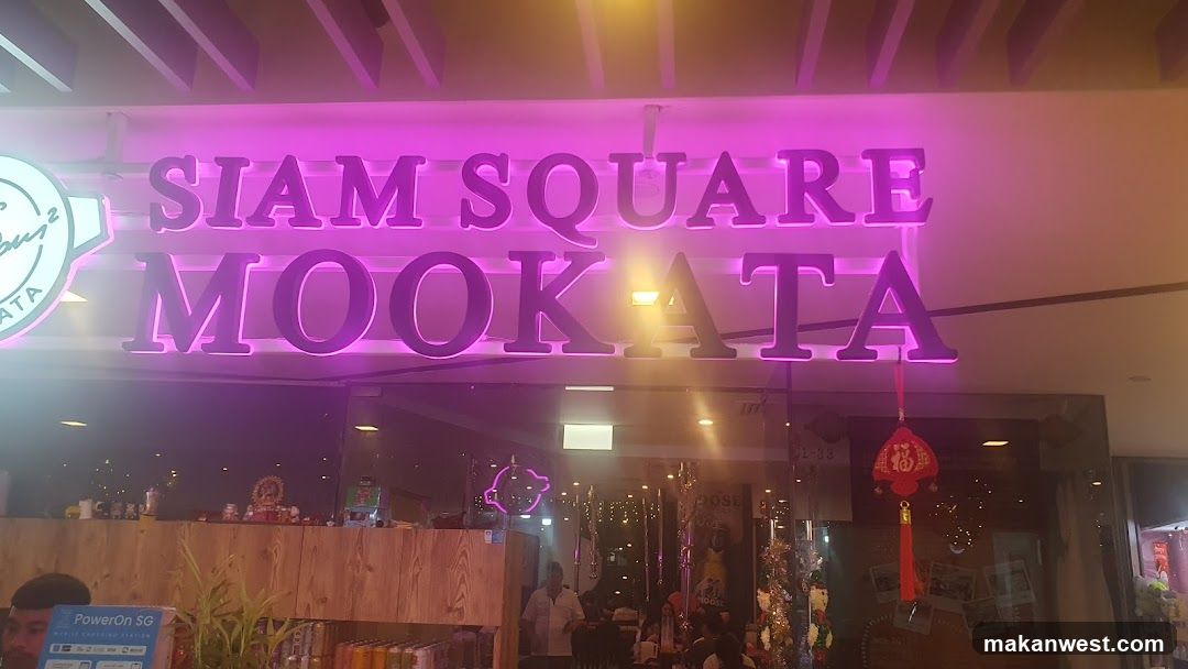 A brightly lit storefront with a large purple neon sign reading 'SIAM SQUARE MOOKATA'. The entrance features glass doors and windows, decorated with hanging ornaments including red Chinese-style lanterns. Inside, there are wooden counters, plants, and people seated at tables, suggesting a casual dining or bar atmosphere.