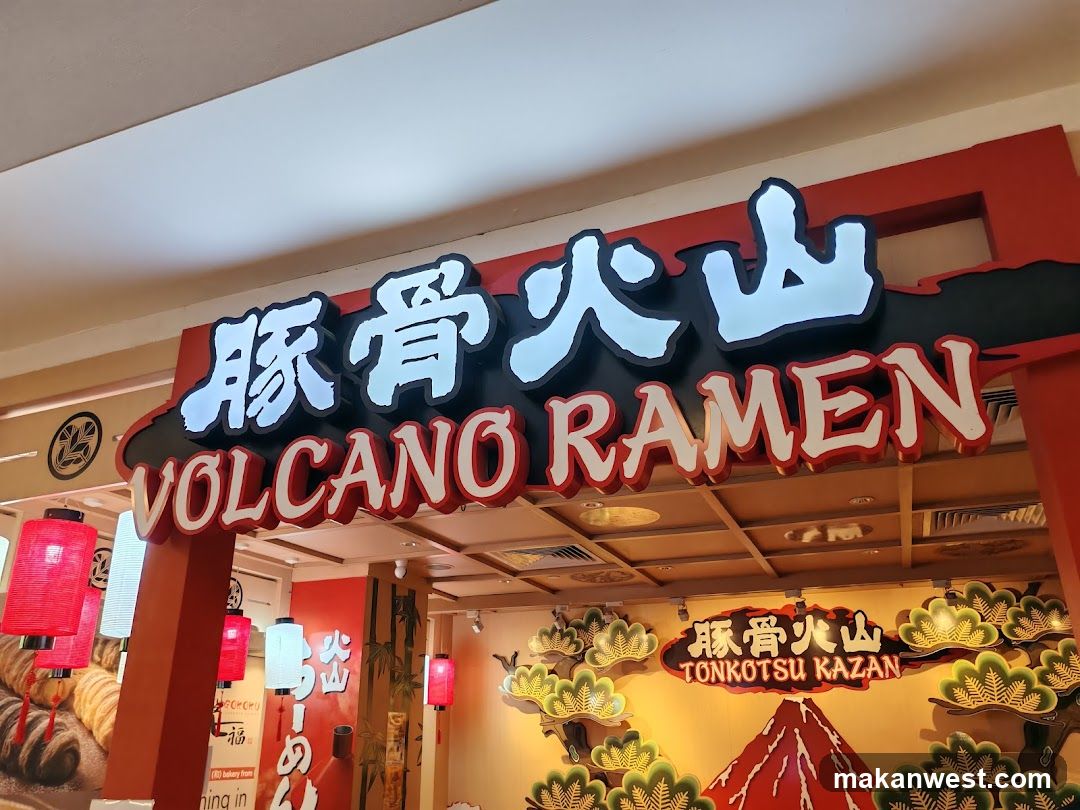 A storefront with a large sign displaying '豚骨火山' and 'VOLCANO RAMEN'. The entrance features red lanterns, decorative elements resembling pine trees and a volcano, and additional signage on the wall inside.