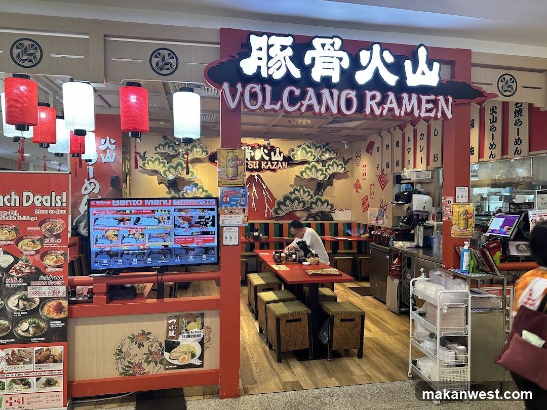 The image shows the storefront of a Japanese ramen shop named 'Volcano Ramen' (豚骨火山). The entrance features a prominent red and white sign with illuminated characters and English text. Red lanterns hang above, and there is a digital menu board displaying various dishes. Inside, tables are set up with stools, and staff can be seen working behind the counter. Decorative elements include stylized trees and traditional Japanese motifs on the walls.
