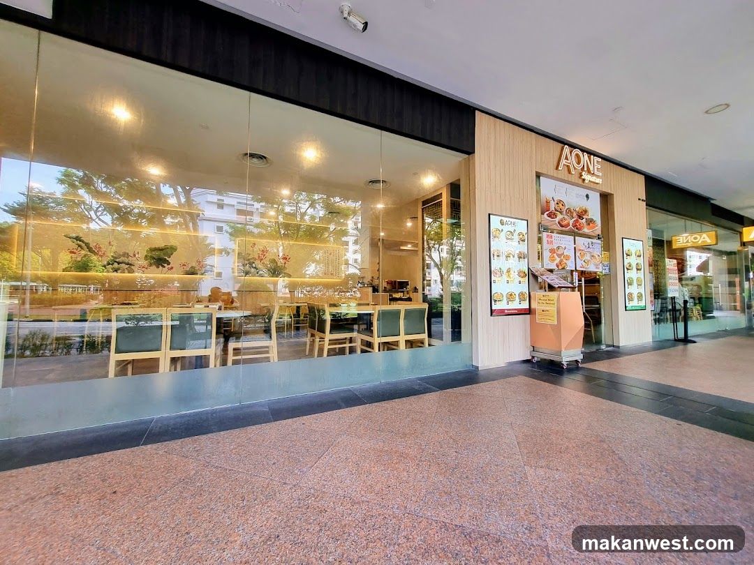 The photo shows the storefront of a restaurant named 'A ONE', featuring large glass windows revealing an interior with wooden tables and chairs. The entrance is flanked by vertical menu boards displaying various food items. A small cart stands near the door, and the flooring outside is tiled in a speckled pattern.