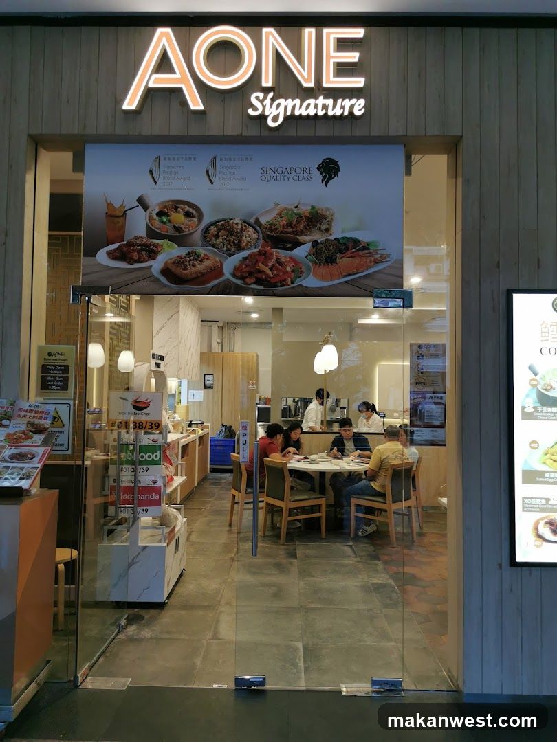 The image shows the storefront of AONE Signature, a restaurant with illuminated signage above the entrance. The facade features wooden paneling and glass doors leading into the dining area where customers are seated at tables. Inside, staff members can be seen attending to guests. A large poster displays various dishes under 'Singapore Quality Class' branding.