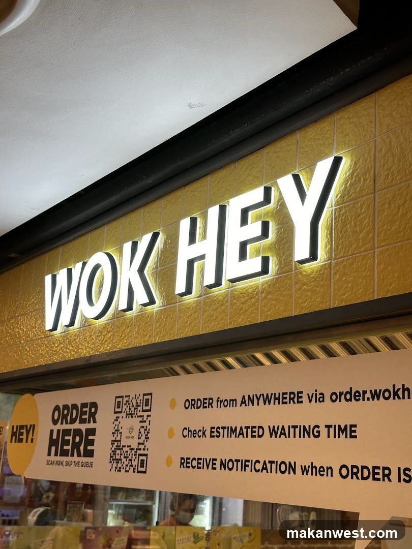 A close-up view of the illuminated sign 'WOK HEY' mounted on a yellow tiled wall above an entrance. Below, a banner promotes ordering via QR code with options to skip queue and receive notifications.