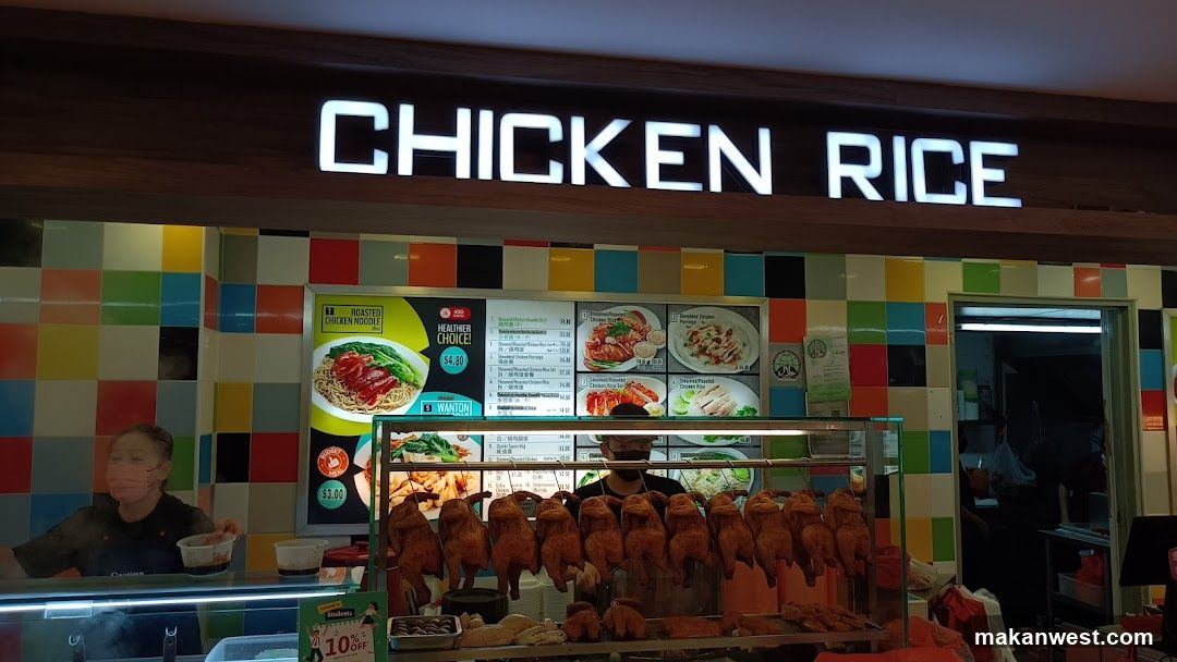 A brightly lit storefront with a large illuminated sign reading 'CHICKEN RICE'. The wall features colorful checkered tiles in yellow, blue, green, and red. Below the sign is a glass display case filled with roasted chickens hanging on hooks. To the left of the display, there are posters advertising food items like 'Roasted Chicken Noodles' and 'Wonton', along with price lists showing prices such as '$4.90' and '$3.10'. A staff member wearing a mask is visible behind the counter on the left side.