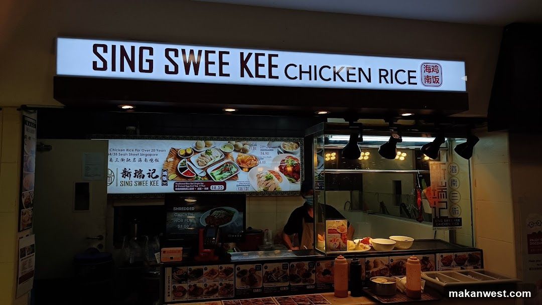 A brightly lit storefront of Sing Swee Kee Chicken Rice, featuring a prominent illuminated sign with Chinese characters and English text. Below the sign, a menu board displays various food items including chicken rice dishes with prices in SGD. The shop interior is visible through glass windows, showing staff preparing food behind the counter. Condiments like soy sauce bottles are placed on the front counter.