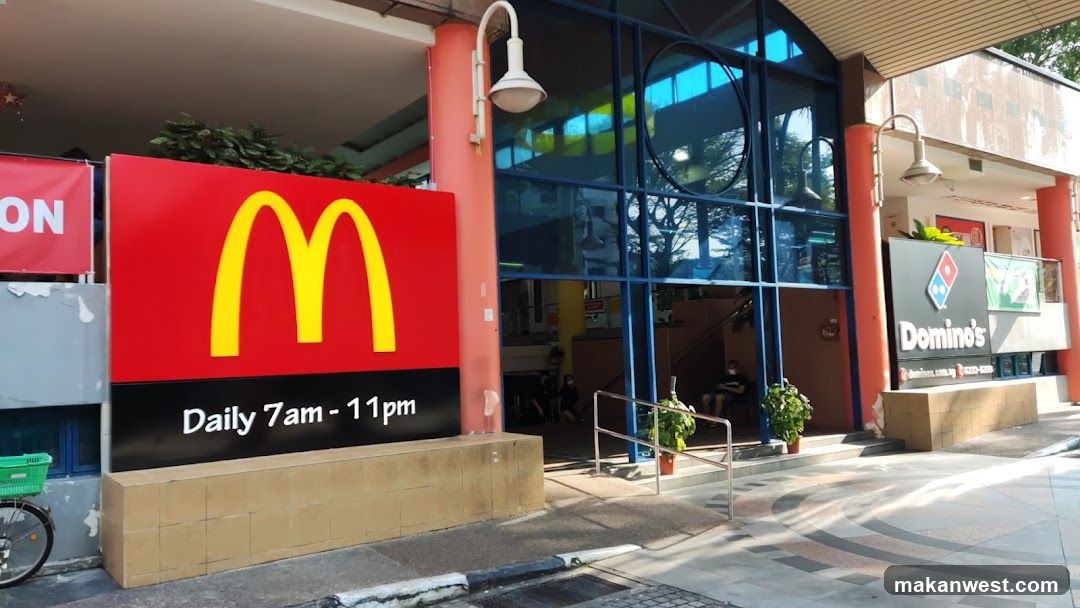 The image shows the exterior of a commercial building housing two fast food chains. On the left, there is a large red sign with the yellow McDonald’s 'M' logo and operating hours listed as Daily 7am - 11pm. To the right, a Domino’s Pizza sign is visible on another part of the building facade. The entrance to both establishments appears accessible via ramps and stairs, with glass doors leading inside.