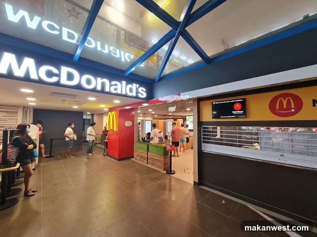 Interior view of a McDonald's restaurant showing customers waiting in line, illuminated signage with 'McDonald's' logo and star design above entrance, red pillars with golden arches, digital screen displaying promotional message, service counter area visible on right side.