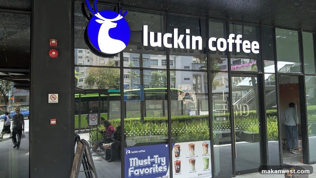 Photo of the exterior entrance and storefront facade of a luckin coffee shop. The building features large glass windows with the brand logo (blue circle with white deer head) and 'luckin coffee' signage mounted above the entrance. A person is visible inside near the door, another sits outside on a bench, and a green bus passes by in the background.