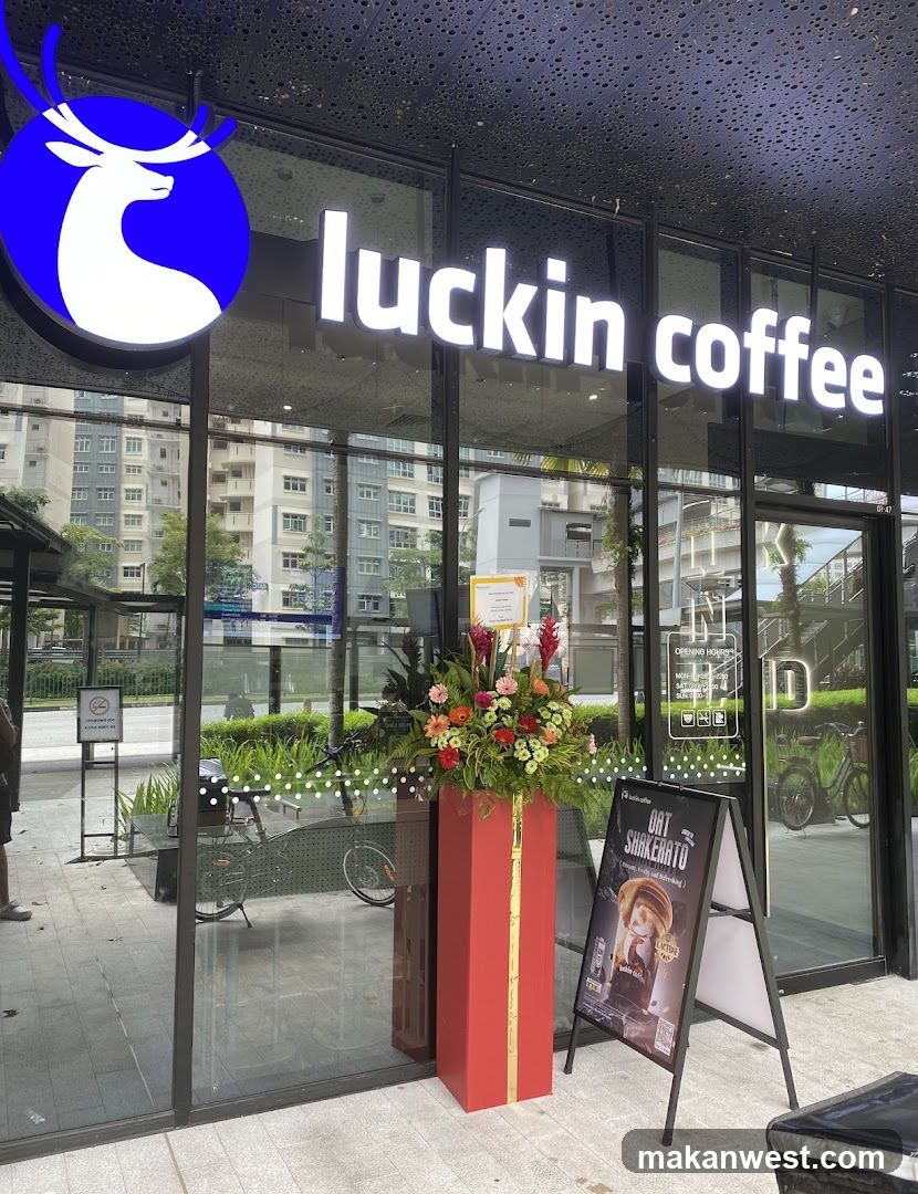 A Luckin Coffee shop entrance with a large illuminated sign and logo on the glass facade. A red floral arrangement stands at the doorway, accompanied by an A-frame promotional board featuring a character advertisement.