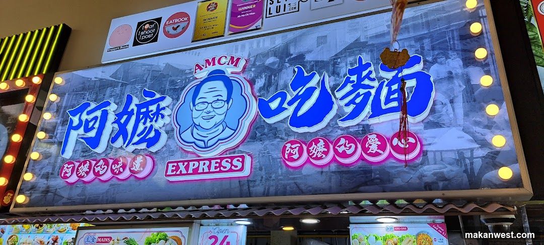 The image shows the exterior of a shop named 'Ama Express' (阿嬤吃麵), featuring a large illuminated signboard with Chinese characters and a cartoon character logo. The sign includes phrases like 'Ama's Taste' and 'Ama's Love'. Above the main sign, there are smaller logos including 'Eatbook', 'Winner', and others. Below the main sign, parts of menu boards or advertisements are visible, showing food items and prices.