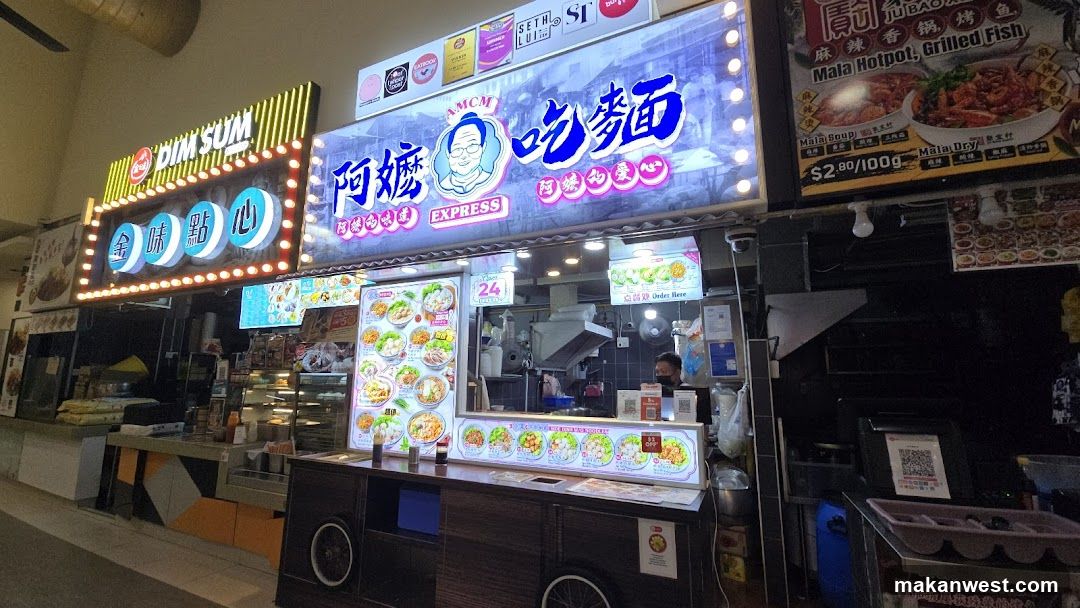 The image shows the exterior of a food stall with bright signage. The main sign reads '阿嬤吃麵' (Auntie's Eat Noodles) and includes an illustration of a woman's face. To the left, another illuminated sign displays '金味點心' (Golden Flavor Dim Sum). Below these signs is a menu board showcasing various noodle dishes with prices. A staff member is visible inside the shop behind the counter.