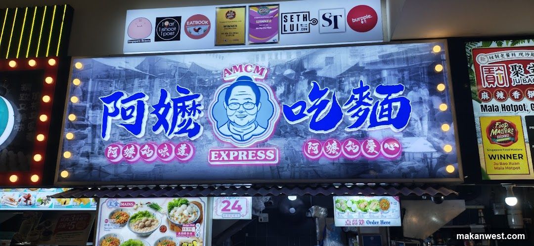 The image shows the exterior of a noodle shop with a large illuminated signboard featuring Chinese characters and an illustration of a smiling face. The sign includes logos from various sponsors like 'Eatbook', 'Seth LUI', and 'burgpole'. Below the main sign, there are smaller signs displaying menu items such as noodles and rice dishes along with prices. The shop appears to be located in a food court or market setting.