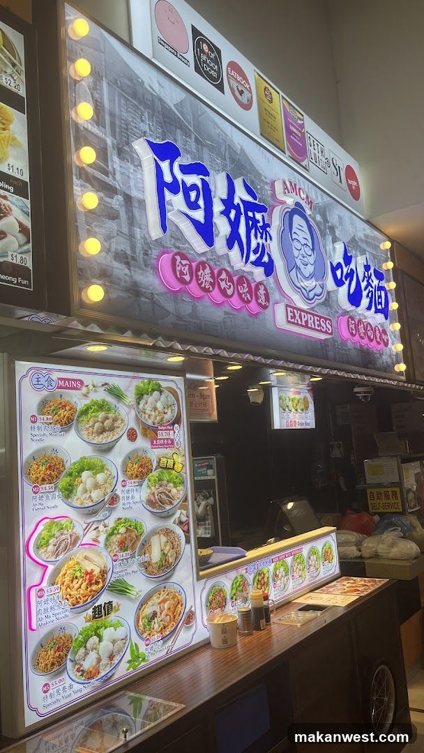The image shows the storefront of a noodle shop with a large illuminated signboard displaying '阿嬤吃麵' (A-Mom Eat Noodles) and 'AMOM EXPRESS'. The sign features a cartoon illustration of an elderly woman's face. Below, there is a menu board showcasing various noodle dishes with prices in dollars. The counter area includes condiments and cups.