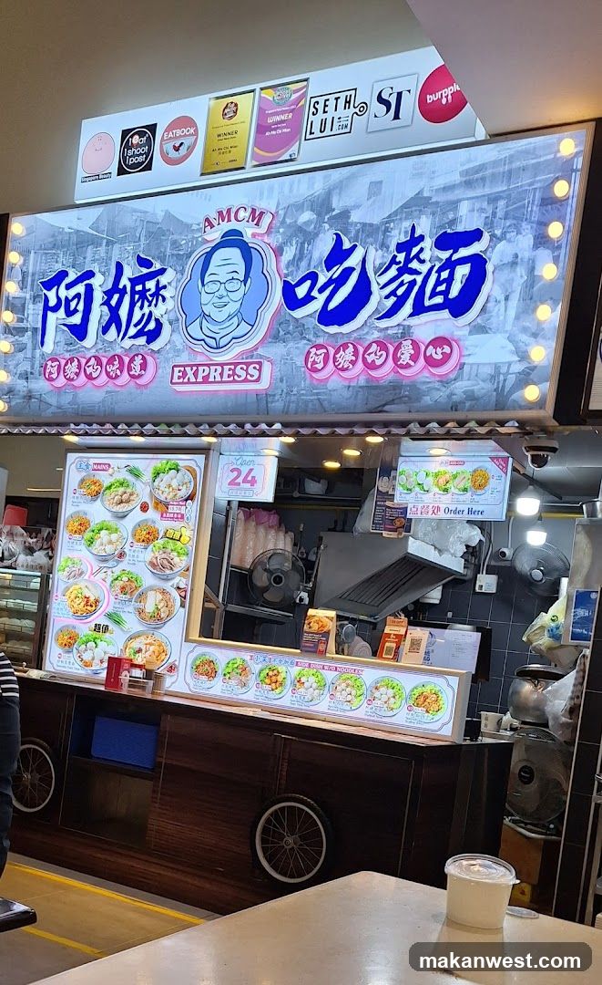 A brightly lit food stall with a large illuminated signboard featuring Chinese characters and an illustration of a smiling man. The sign includes logos from sponsors like Seton, Burger King, and others. Below the sign is a menu board displaying various noodle dishes with prices. The counter area shows cooking equipment, including a fan and exhaust hood. A plastic cup sits on a table in the foreground.
