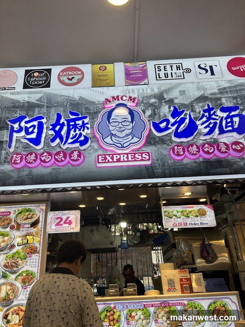 A food stall with a large signboard displaying 'AMCM' and Chinese characters, featuring a cartoon character logo. The shop has visible menu boards on the sides showing various dishes like noodles and rice bowls. A customer is standing in front of the counter.