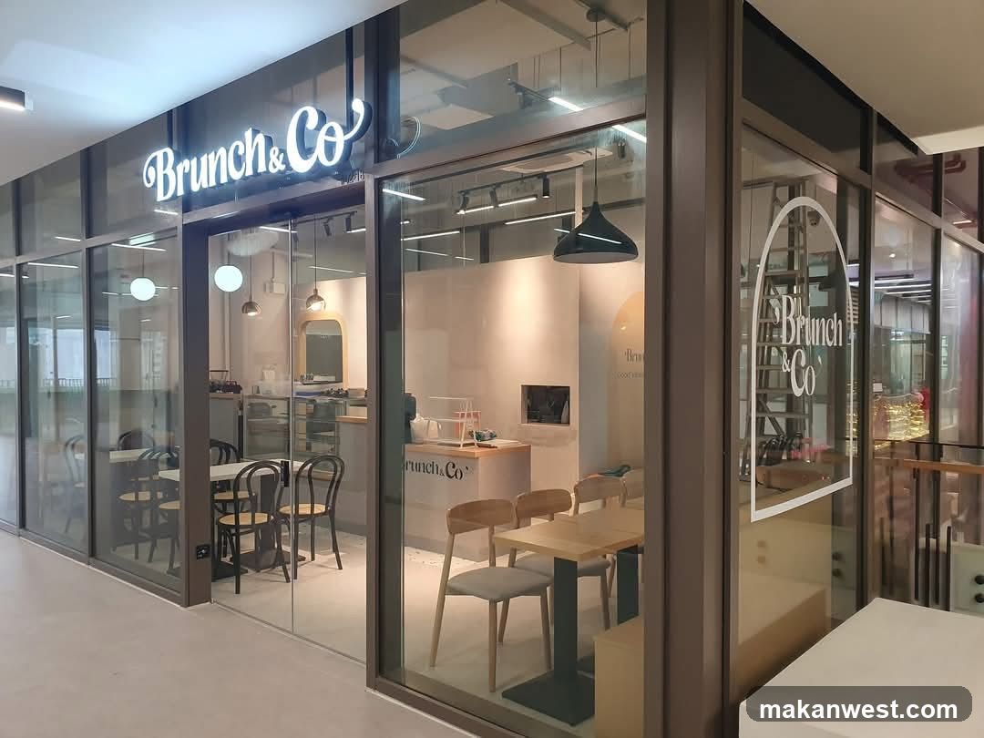 A modern glass-fronted cafe with illuminated 'Brunch & Co' signage above the entrance. Interior shows wooden tables, chairs, and a counter area visible through large windows.