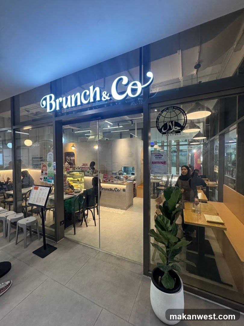 The photo shows the storefront of a cafe named 'Brunch & Co.' with illuminated signage above the entrance. The shop has large glass windows and doors, allowing visibility into the interior where tables are set up for customers. Inside, there is a counter area displaying various food items like pastries or sandwiches on trays. A potted plant sits near the entrance, adding greenery to the scene. There's also a circular logo sticker on one of the glass panels featuring Arabic calligraphy. Outside, some chairs are visible, suggesting an outdoor seating option. The overall ambiance appears modern and inviting.