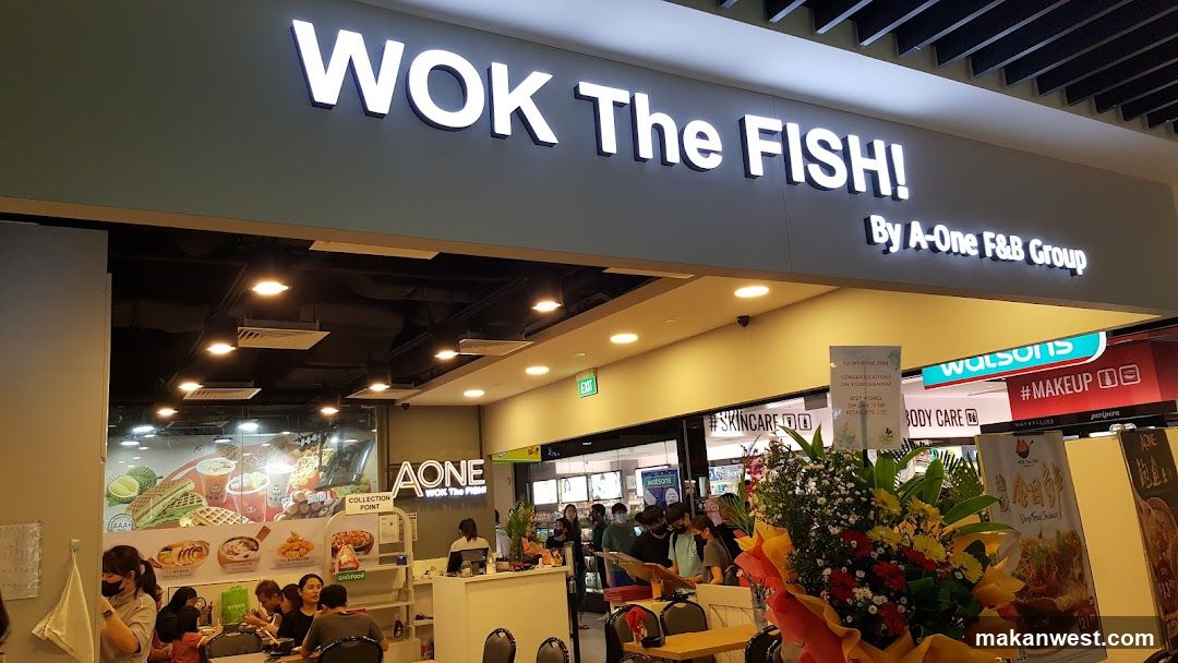 A storefront with a large illuminated sign reading 'WOK The FISH!' and smaller text 'By A-One F&B Group'. Inside, customers are seated at tables near the entrance. A floral arrangement is visible in the foreground.