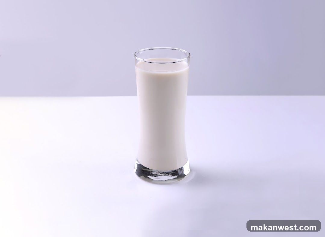 A tall, clear glass filled with white milk sits on a white surface against a light purple background.