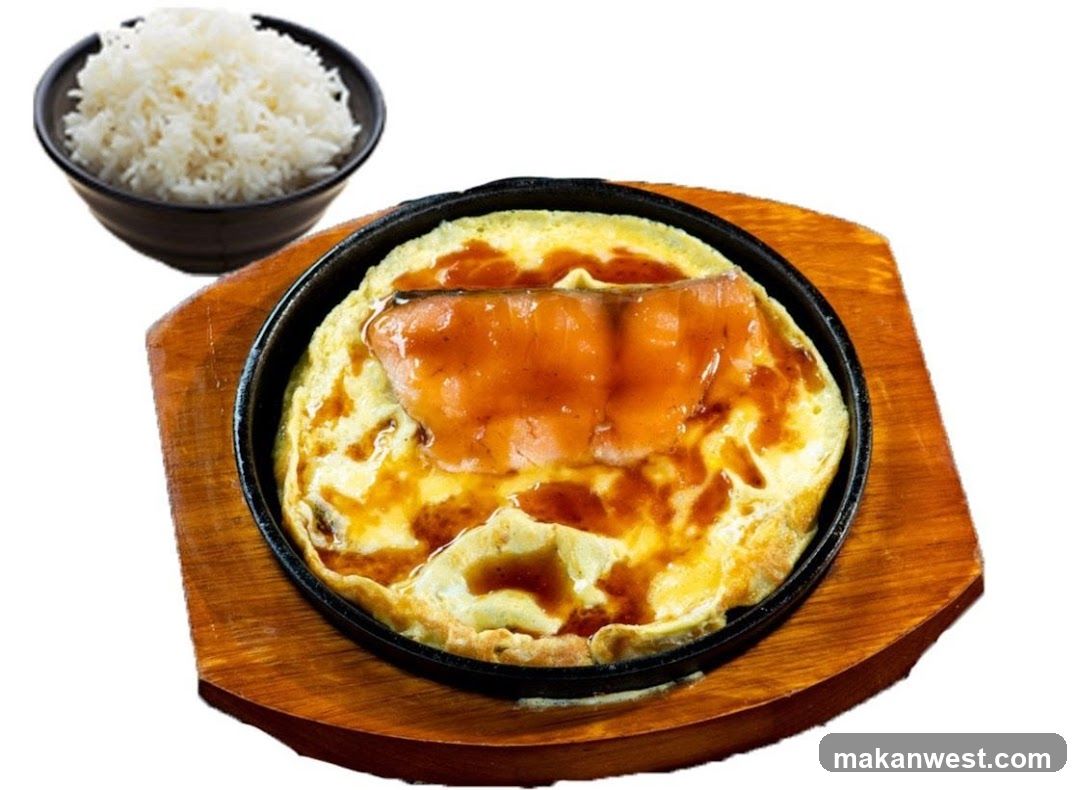A close-up shot of a sizzling dish featuring a piece of salmon glazed with a dark sauce, served over a bed of cooked eggs in a black cast-iron skillet. The skillet rests on a wooden base. To the side, a small black bowl is filled with fluffy white rice. The background is white.